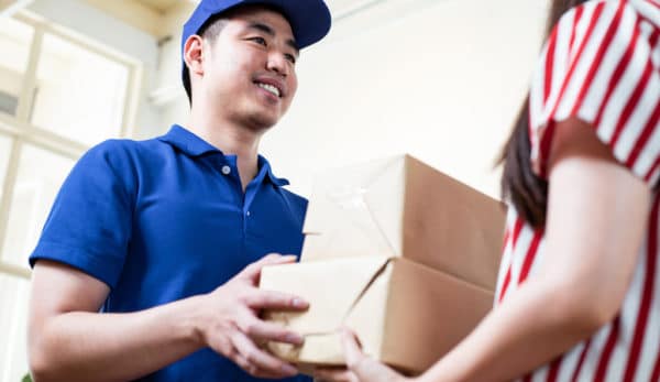 Delivery man giving a cardboard box to young woman at entrance of her home of fast and reliable service. Postman delivers the parcel to girl at home with smile. Delivery and mailing service concept.